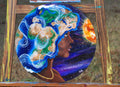 A circular painted artwork leaning against a wooden easel outdoors. The painting shows a side profile of a person with a flowing blue, green, and tan afro in the pattern of earth, set against a starry purple and blue space scene with a bright orange sun.